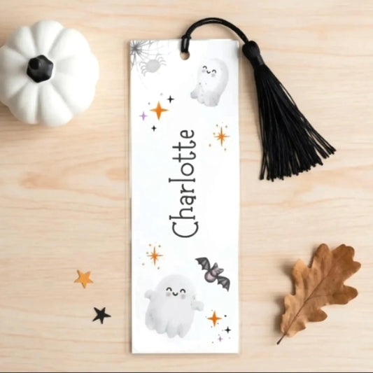 A white bookmark with a black tassel, featuring a spider web design and illustrations of a ghost, bat, and leaf, along with the name "Charlotte" written on it. The bookmark is placed on a wooden surface, accompanied by a small white pumpkin and a brown leaf.