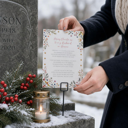 A person is holding a card with a Christmas message in front of a gravestone, with a lit candle and berries nearby.