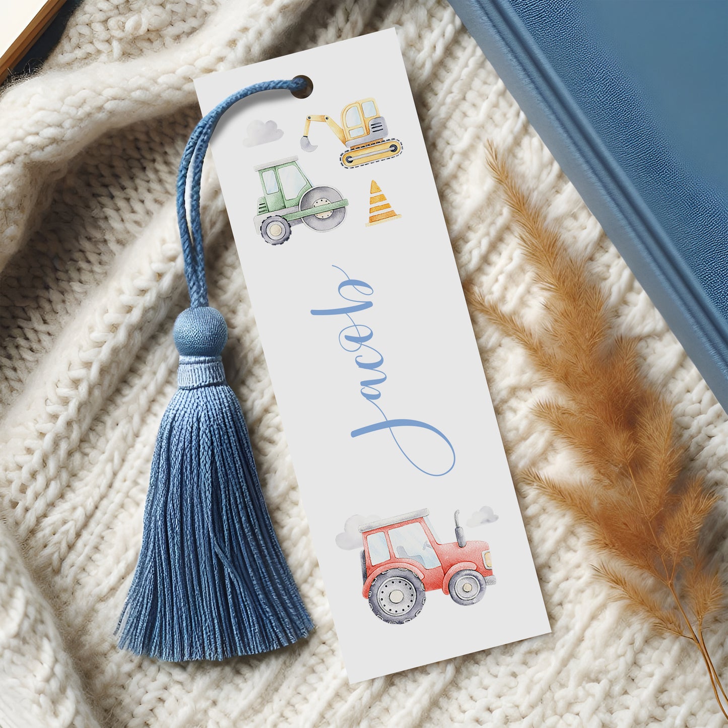 A white bookmark with illustrations of construction vehicles and a blue tassel, placed on a white knitted blanket.