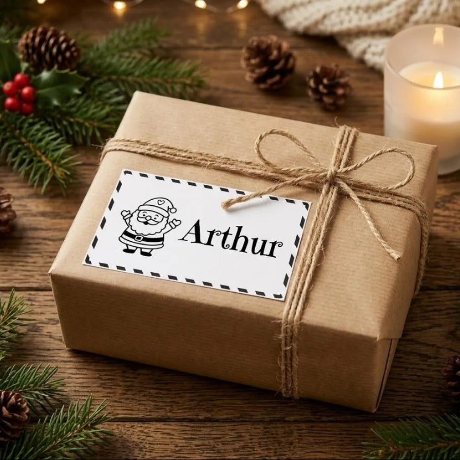 A gift box with a Santa Claus label, surrounded by pine branches, pine cones, and a lit candle on a wooden surface.