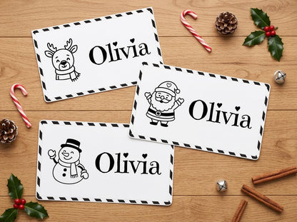 three personalized nameplates with illustrations of Santa Claus and a snowman, along with the names "Olivia" written on them. The nameplates are placed on a wooden surface, surrounded by holiday decorations such as candy canes, pine cones, and holly leaves.
