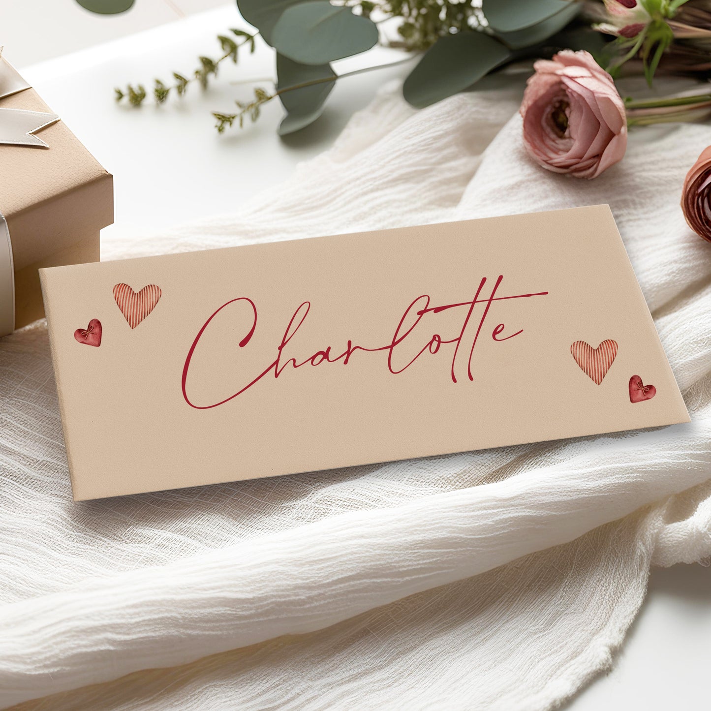 a beige card with the name "Charlotte" written on it, surrounded by small red hearts. The card is placed on a white cloth, and there are some pink flowers and a gift box in the background.