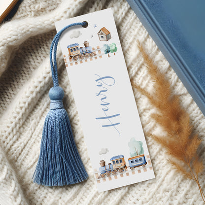 A white bookmark with a blue tassel and a train illustration on it, placed on a white knitted blanket.