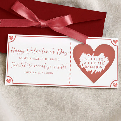 a Valentine's Day gift card with a red ribbon and a heart-shaped balloon design. The card is placed on a beige cloth, and the text on the card reads "Happy Valentine's Day to my amazing husband. Scratch to reveal your gift! Love, Erika XOXOXO".