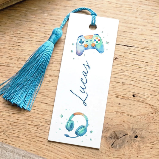 A white bookmark with a blue tassel and a gaming controller design, along with a name tag, is placed on a wooden surface.