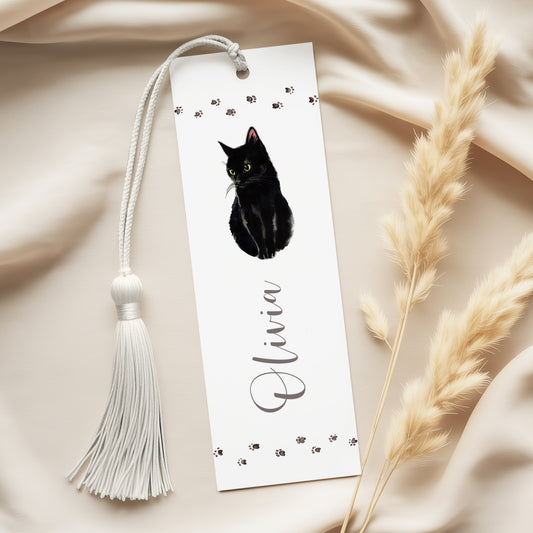A white bookmark with a black cat illustration and the name "Olivia" written on it, along with a white tassel. The bookmark is placed on a beige fabric surface, with some dried plant material visible to the right.