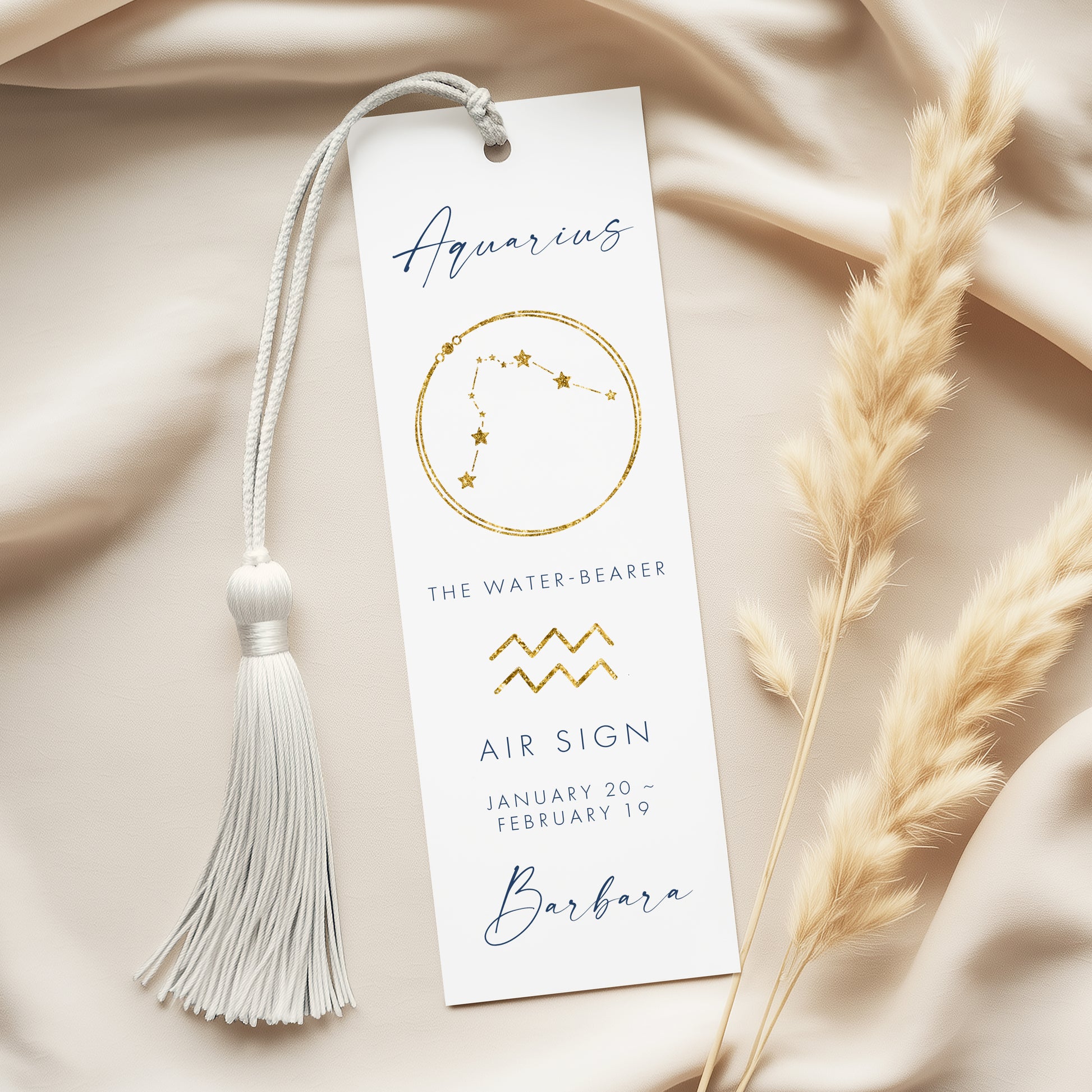 a white bookmark with the name "Barbara" printed on it, along with the astrological sign "Aquarius" and the zodiac symbol for the month of January. The bookmark is placed on a beige fabric, with a white tassel hanging from the top.