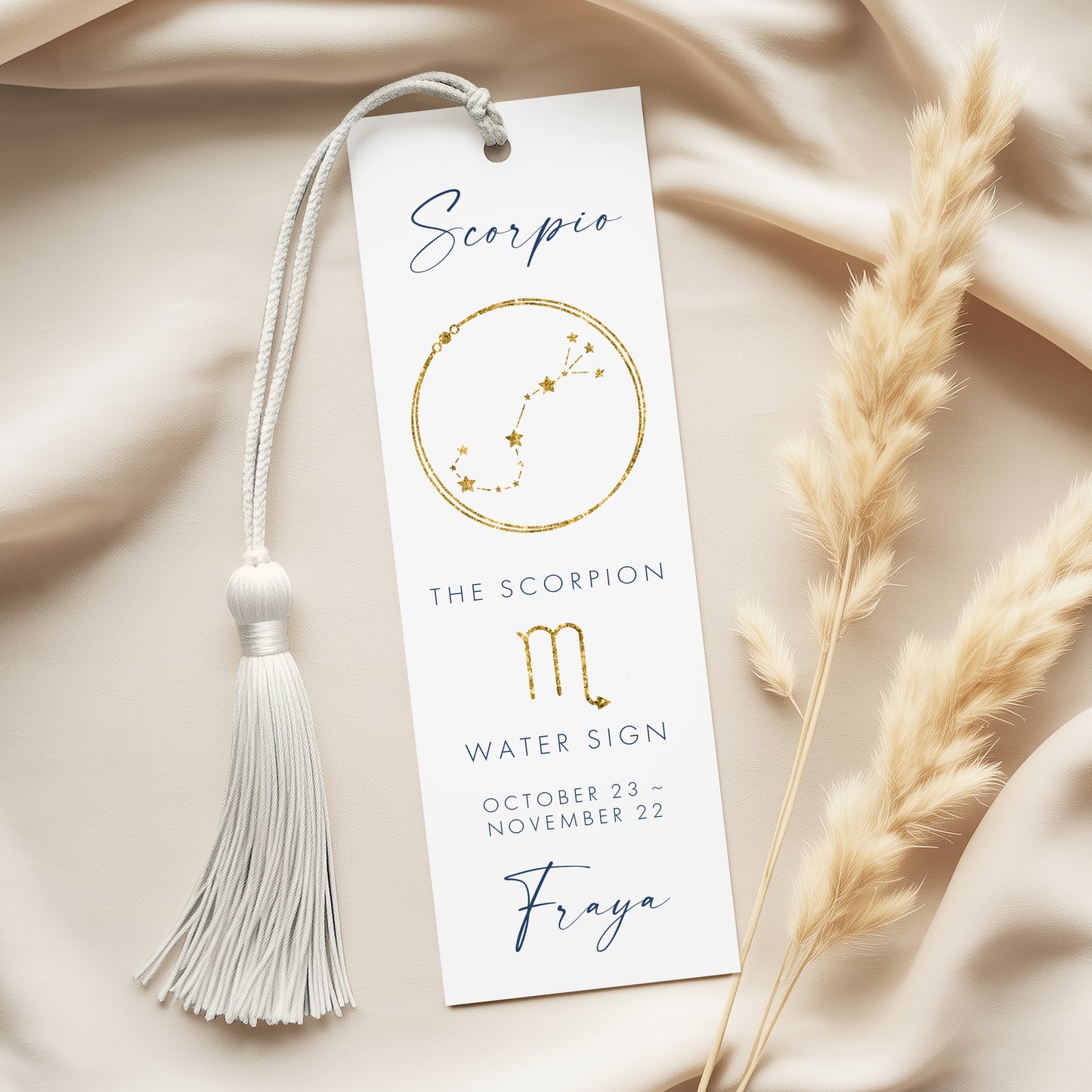 A white bookmark with a tassel and a design of the Scorpio zodiac sign, along with some dried reeds, placed on a beige fabric background.