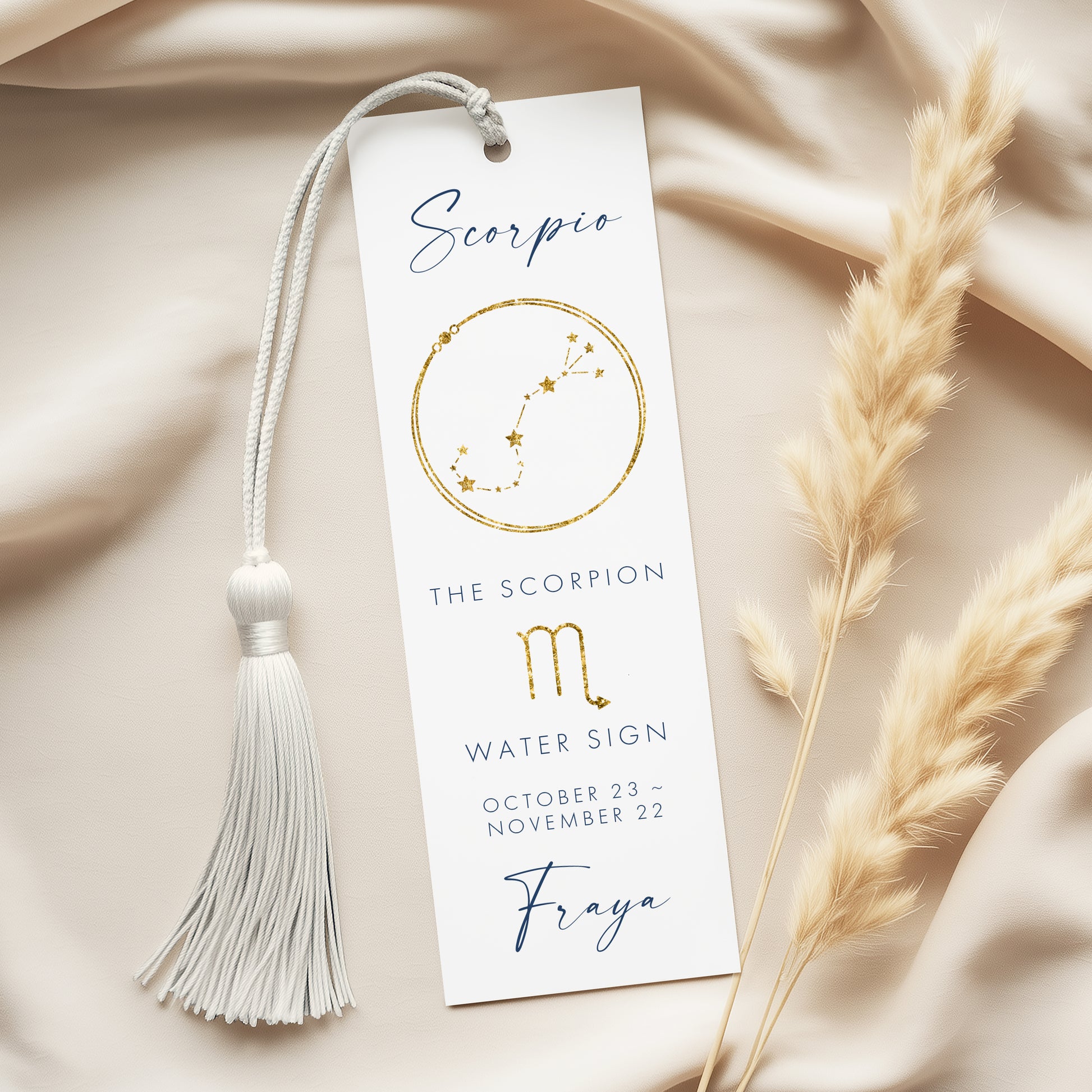 A white bookmark with a tassel and a design of the Scorpio zodiac sign, along with some dried reeds, placed on a beige fabric background.