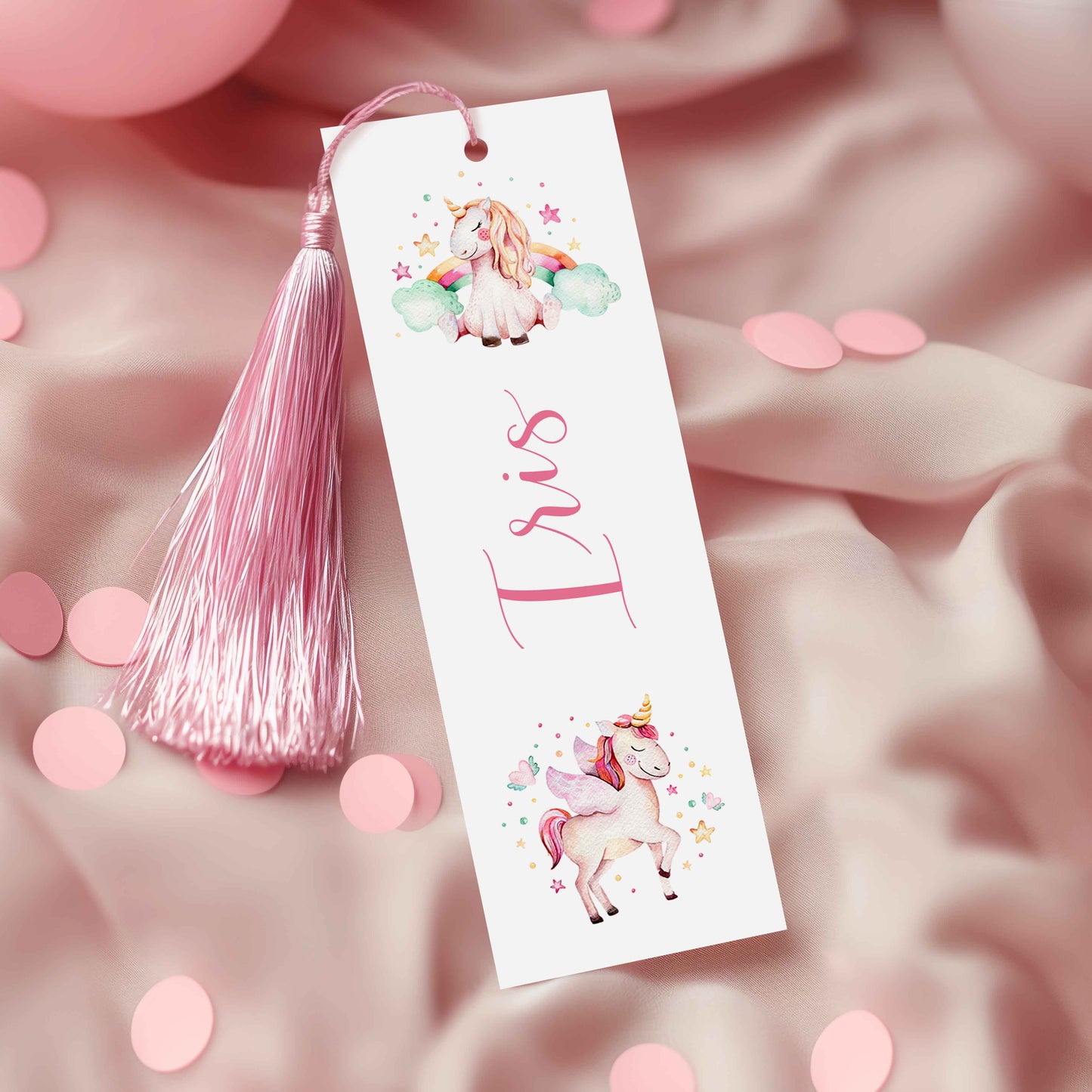 A white bookmark with a pink tassel and illustrations of a unicorn and a fairy on it, placed on a pink fabric background with scattered confetti.