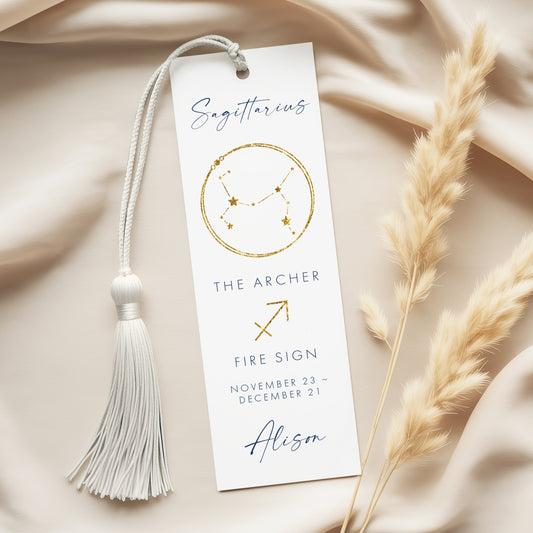 a white bookmark with a tassel and a gold-colored astrological symbol, placed on a beige fabric background.