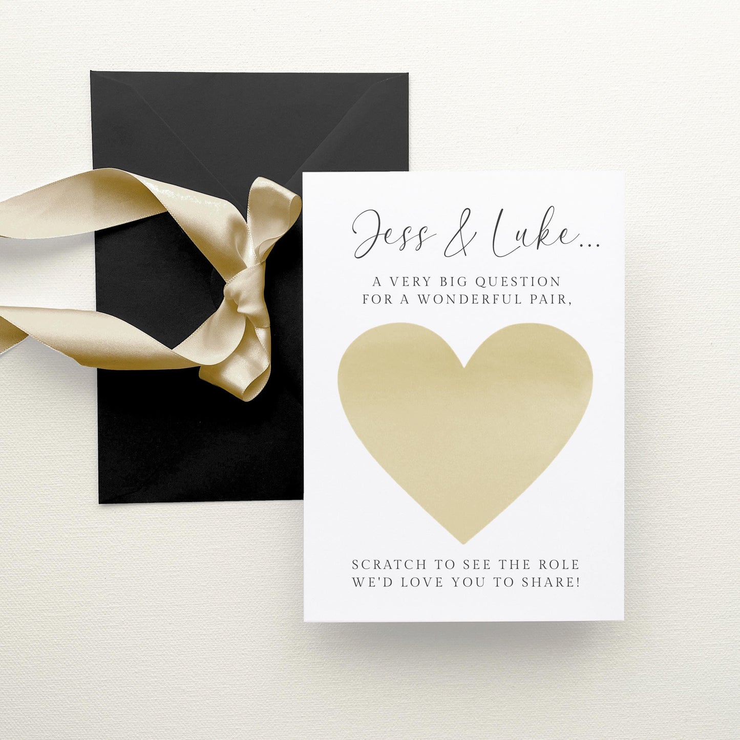 A white card with a gold heart-shaped scratch-off, accompanied by a black envelope with a gold ribbon.
