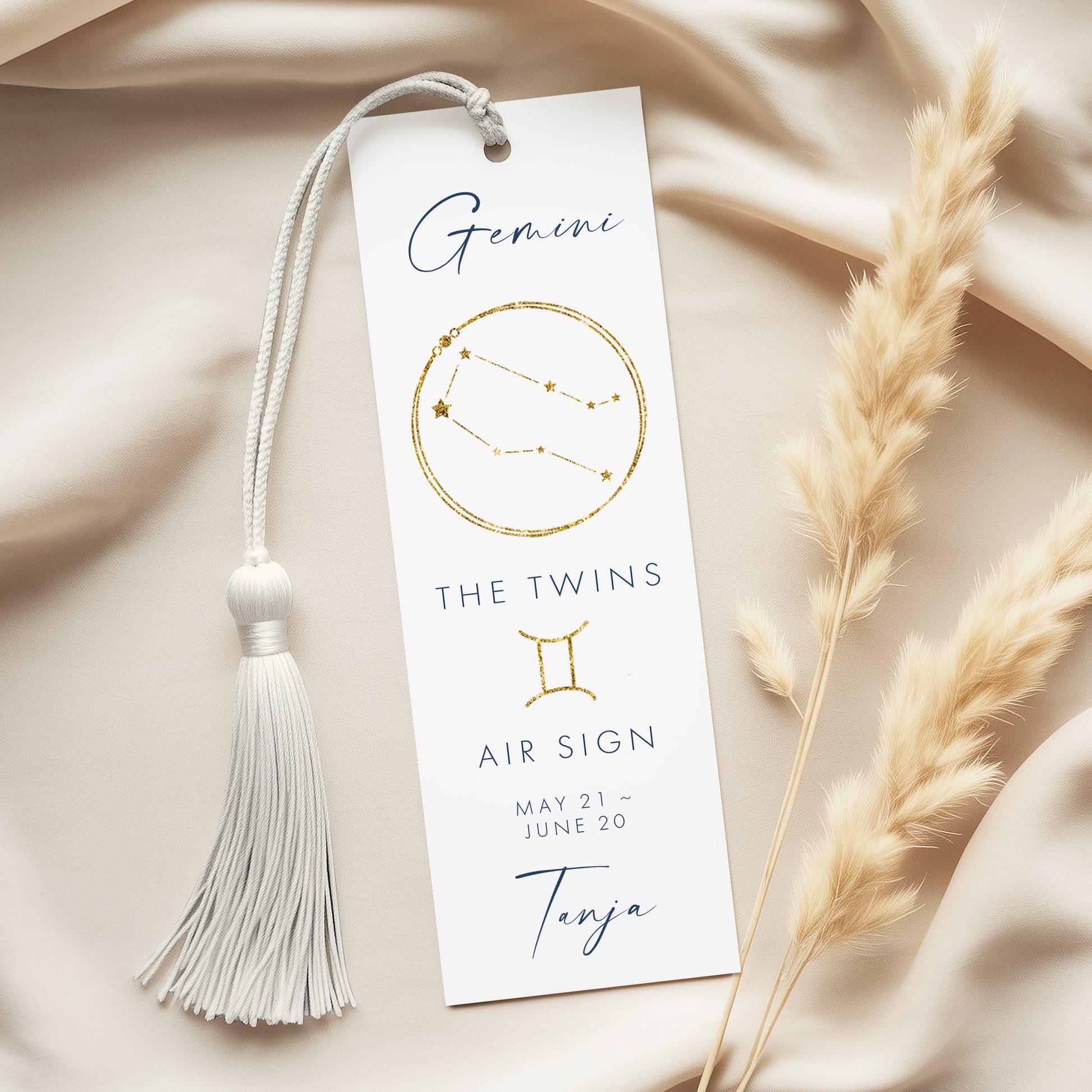 a white bookmark with a tassel and a gold-colored zodiac symbol, placed on a beige fabric background.