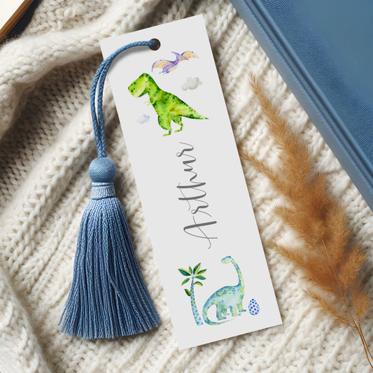 A white bookmark with a blue tassel and illustrations of a green dinosaur and a blue dinosaur on a white background.