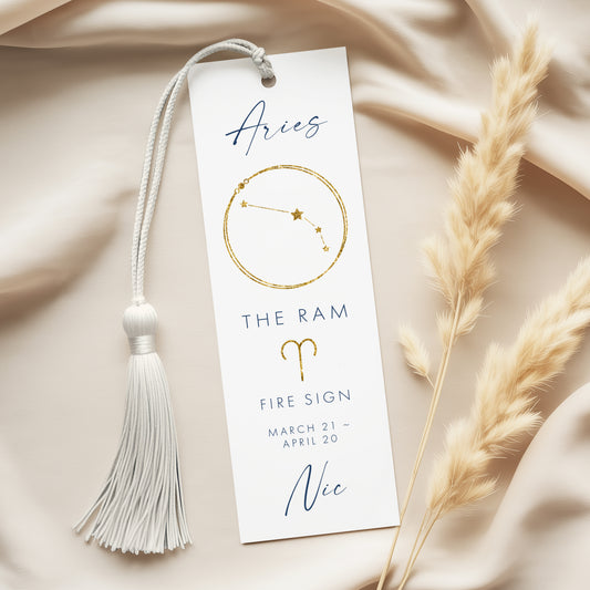 a white bookmark with a tassel and a gold-colored astrological symbol, placed on a beige fabric background.