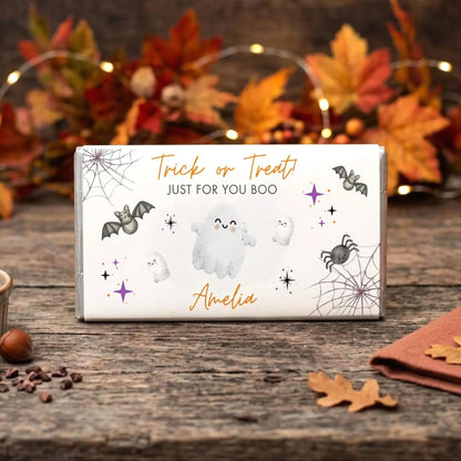 a Halloween-themed candy bar with a white wrapper, decorated with bats, ghostly figures, and autumn leaves. The candy bar is placed on a wooden surface, surrounded by various Halloween-themed items such as a bowl of nuts, a brown napkin, and some fallen leaves.