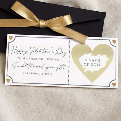 a Valentine's Day gift card with a heart-shaped design and a gold ribbon. The card is placed on a beige surface and has a black envelope tied with a gold ribbon on top.