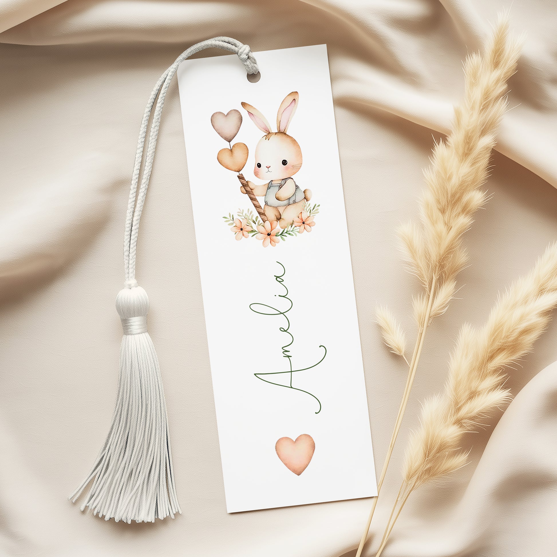 A white bookmark with a tassel and a small bunny illustration on it, placed on a beige fabric background.