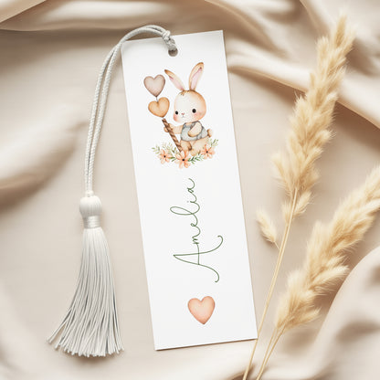 A white bookmark with a tassel and a small bunny illustration on it, placed on a beige fabric background.