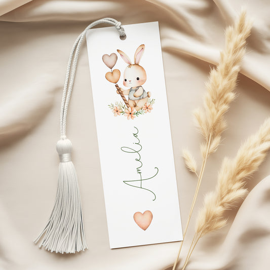 A white bookmark with a tassel and a small bunny illustration on it, placed on a beige fabric background.